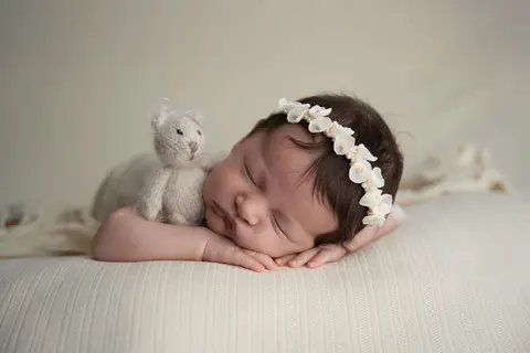 Новородено с бели цветчета спи върху ръчичките си, а до него се сгушва пухкаво мече