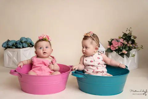 Близначки с цветя в косите седят в две метални коритца, розово и тюркоазено