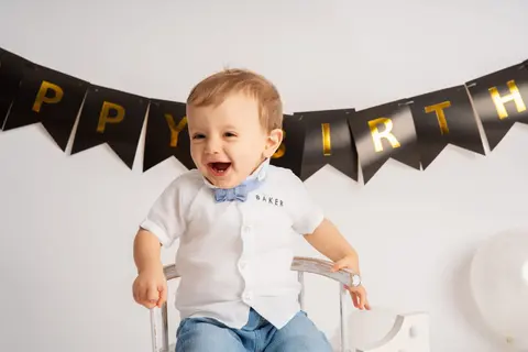 Момченце в бяла риза се смее с глас под черно-златен банер Happy Birthday