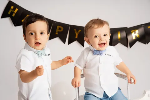 Две момченца с папийонки, едното учудено, другото засмяно, под банер Happy Birthday
