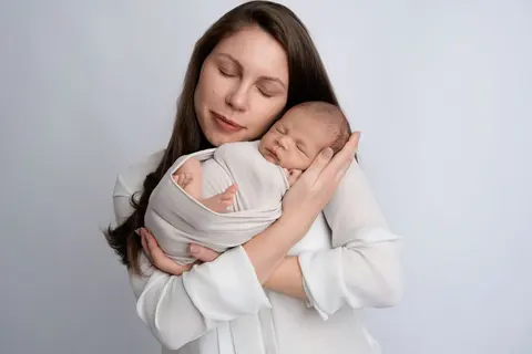 Мама със затворени очи притиска нежно бебето до бузата си, облечена в бяло