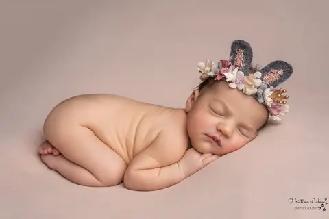 Новородено с лента със заешки ушички и цветя спи свито на коремче на пудров фон