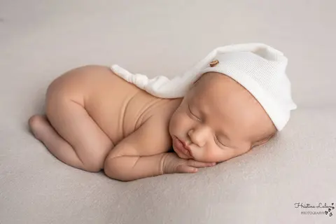 Новородено с бяла нощна шапчица спи свито на коремче с ръчички под бузката