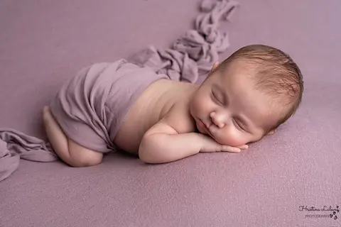 Новородено спи по коремче, наполовина завито с лилав плат, на люляков фон